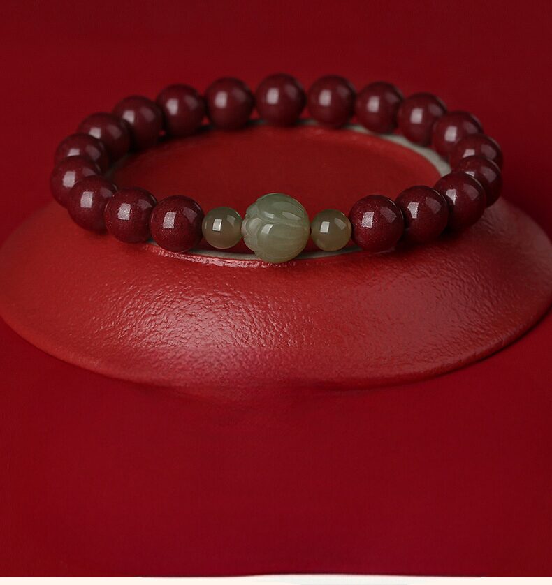 A single-strand bracelet of nineteen deep red polished cinnabar beads with a carved pale green Hetian jade lotus focal bead and two flanking jade rounds, on a dark background.