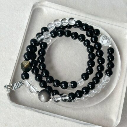 Triple-wrap black obsidian and white crystal bracelet with silver koi fish charm, three-strand construction, volcanic black and ice clear crystal, silver obsidian focal bead, dark background