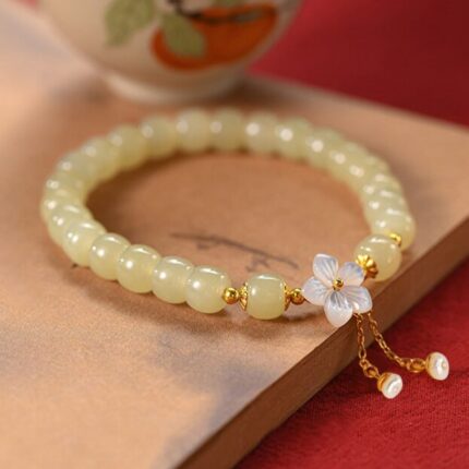 : A single-wrap Hetian jade bracelet in pale green with a white shell flower charm and dangling gold tassel ending in freshwater pearls, on a white background.