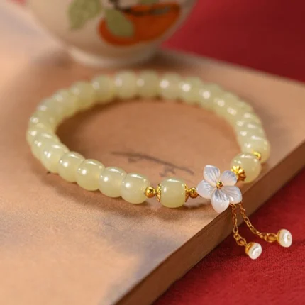 : A single-wrap Hetian jade bracelet in pale green with a white shell flower charm and dangling gold tassel ending in freshwater pearls, on a white background.