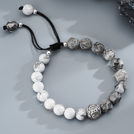 A gradient macrame bracelet shifting from white marble howlite beads to grey map stone beads, centered with an S925 silver Six Syllable Mantra bead, on a dark background.