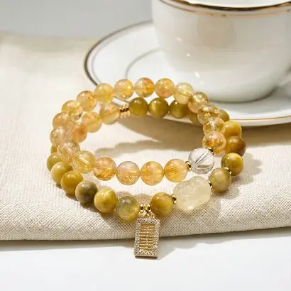 Double-wrap yellow rutilated quartz and golden tiger's eye bracelet laid flat on white linen, with yellow crystal Pixiu carving, clear quartz cube, rhinestone abacus pendant, warm golden tones