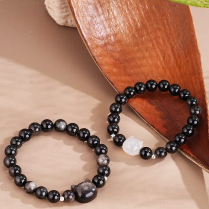 Both bracelets laid flat side by side on a neutral background — silver obsidian cat head version on the left, white agate cat head version on the right — showing the cat face detail and obsidian bead quality clearly.