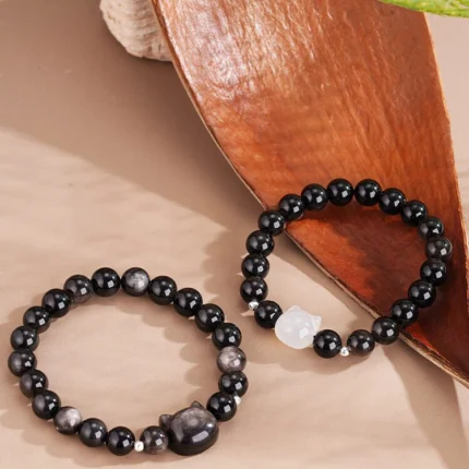 Both bracelets laid flat side by side on a neutral background — silver obsidian cat head version on the left, white agate cat head version on the right — showing the cat face detail and obsidian bead quality clearly.