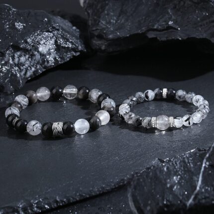 A pair of matching Black Obsidian and Black Rutilated Quartz elastic stretch bracelets, shown side by side on a white background. The larger men's bracelet features 10 mm beads with a textured S925 silver ring accent. The smaller women's bracelet has 8 mm beads with twin silver ring details. A size label sits between the two pieces.