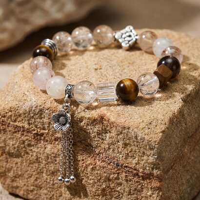 Black Gold Super Seven multi-gem bracelet laid flat on white linen, showing black and gold thread inclusions, tiger's eye beads, ice obsidian and white crystal cube, zinc alloy tassel