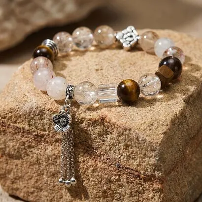 Black Gold Super Seven multi-gem bracelet laid flat on white linen, showing black and gold thread inclusions, tiger's eye beads, ice obsidian and white crystal cube, zinc alloy tassel